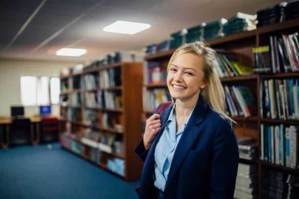 A young woman standing in a library surrounded by bookshelves while applying for student admissions online.