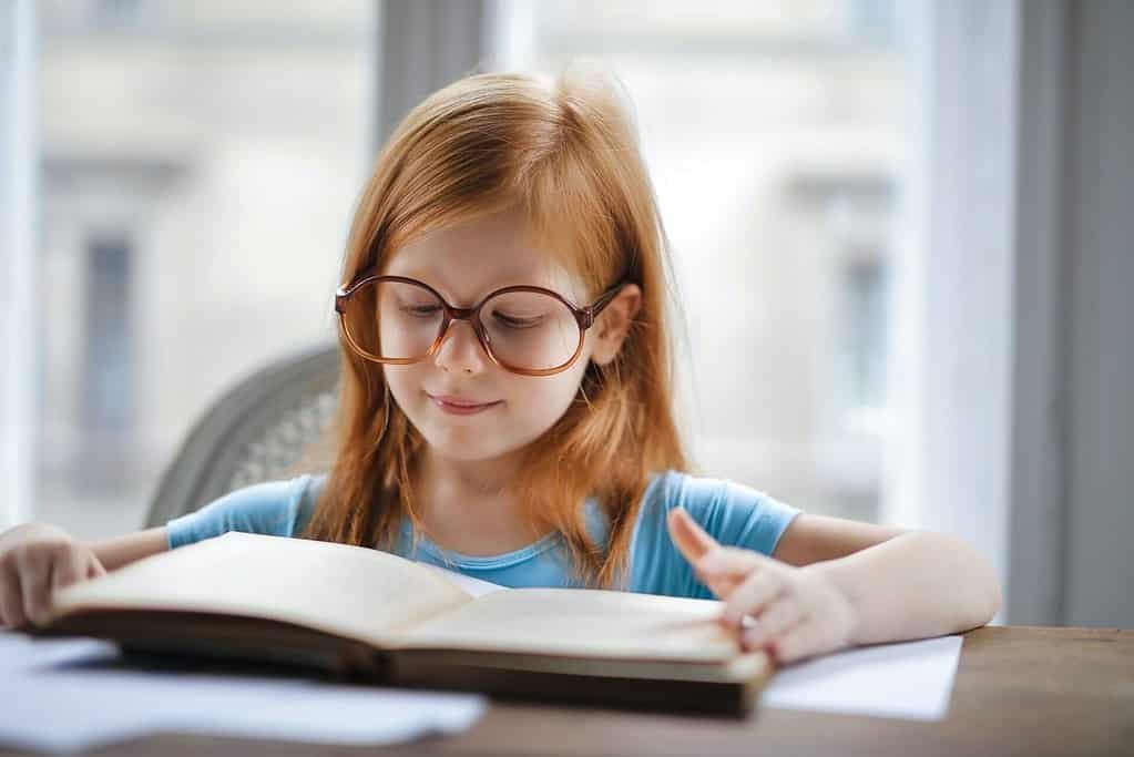 A little girl wearing glasses is reading a book, showcasing talent.